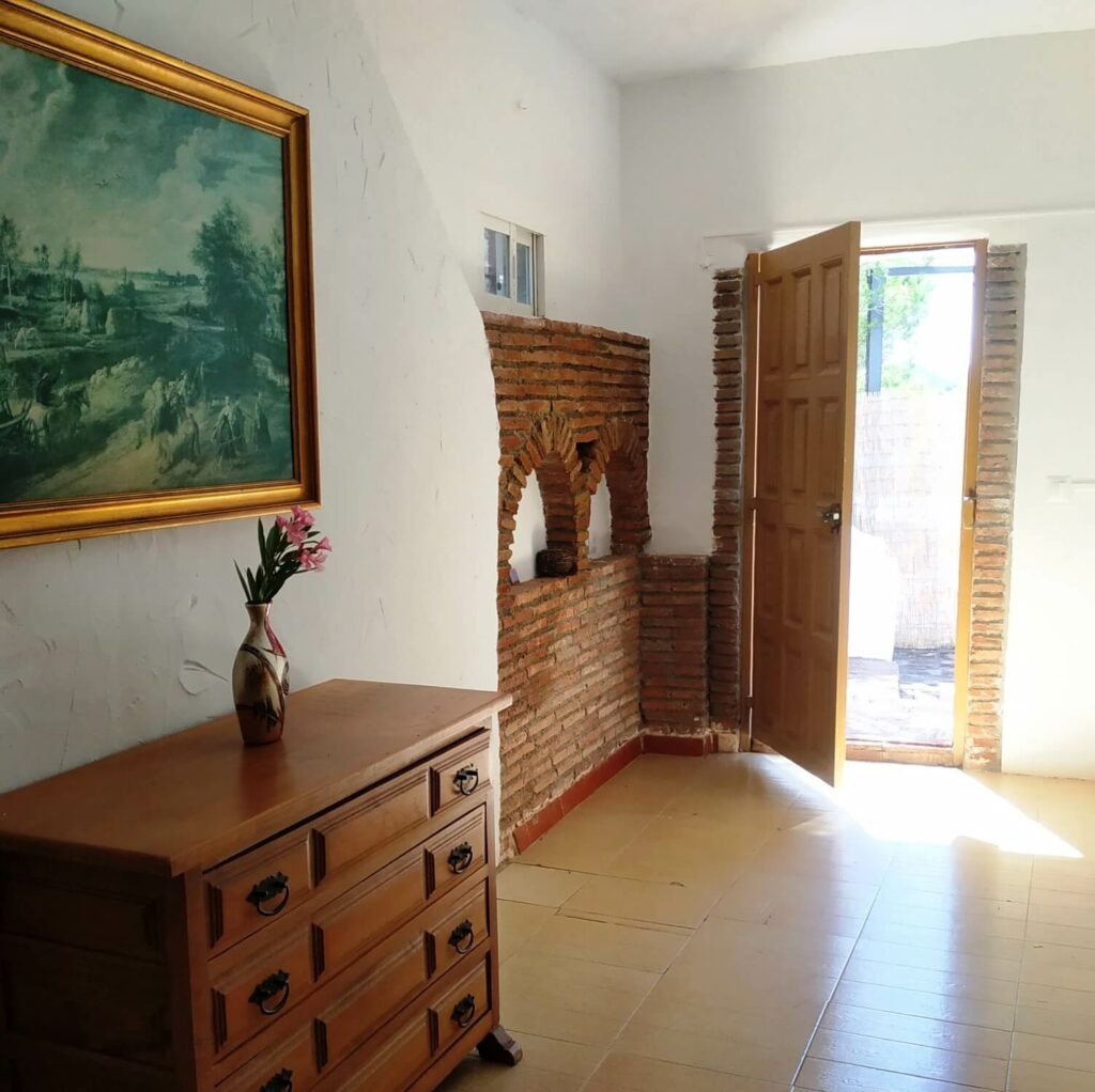 Entrada de una casa vista desde dentro con la puerta abierta. A la izquierda, en primer plano, una gran cómoda coronada por un cuadro.