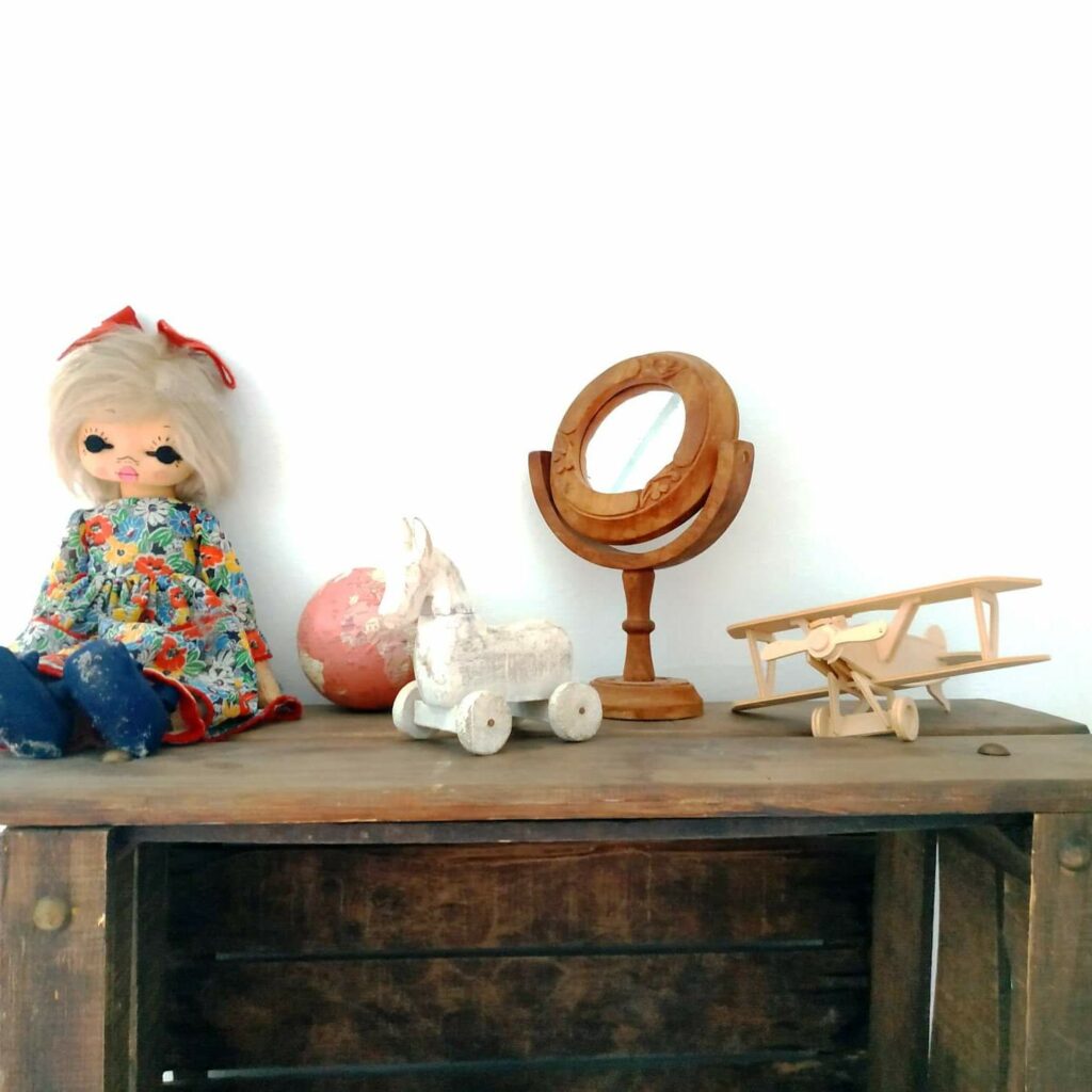 Eine Holzkiste, die an einer weißen Wand befestigt ist und auf der eine alte Puppe, eine Miniaturweltkarte, ein kleines weißes Holzpferd, ein kleiner Spiegel und ein Holzflugzeug liegen.