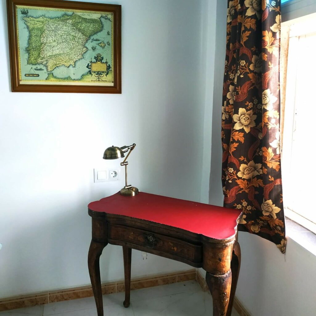 hoek van een kamer met een gordijn met grote bruine bloemen en een oude houten sokkeltafel met een rode deeus, met daarop een antieke bureaulamp