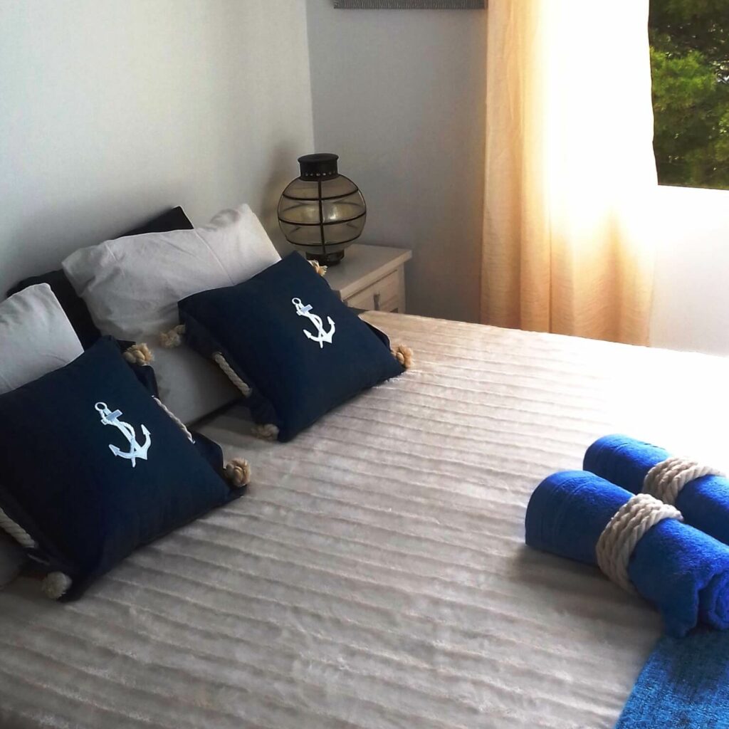 White bed by a window, with navy blue cushions decorated with a sea anchor