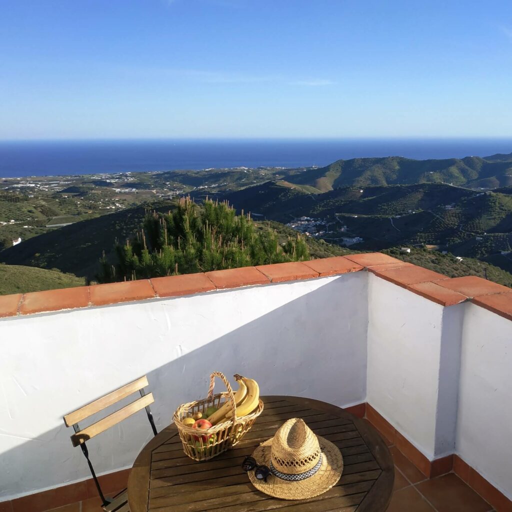 La Méditerrannée et les montagnes de Malaga vues d'un terrasse avec une table sur laquelle sont posés un panier de fruits et un chapeau de paille.
