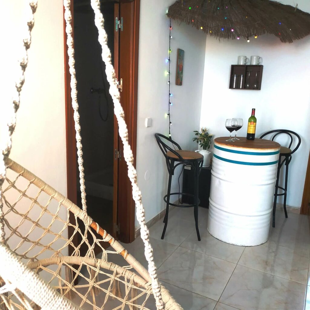 The bar consists of a white metal can with a bottle of red wine and two glasses served on top. In the foreground a white rope chair is suspended.