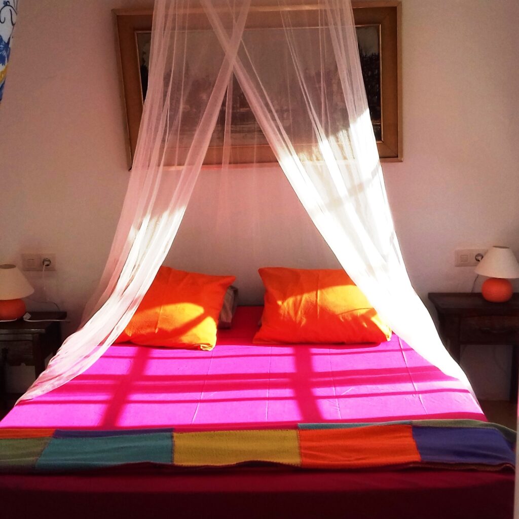 Pink bed topped with a mosquito net seen from the front, with a brightly coloured plaid and orange pillows.