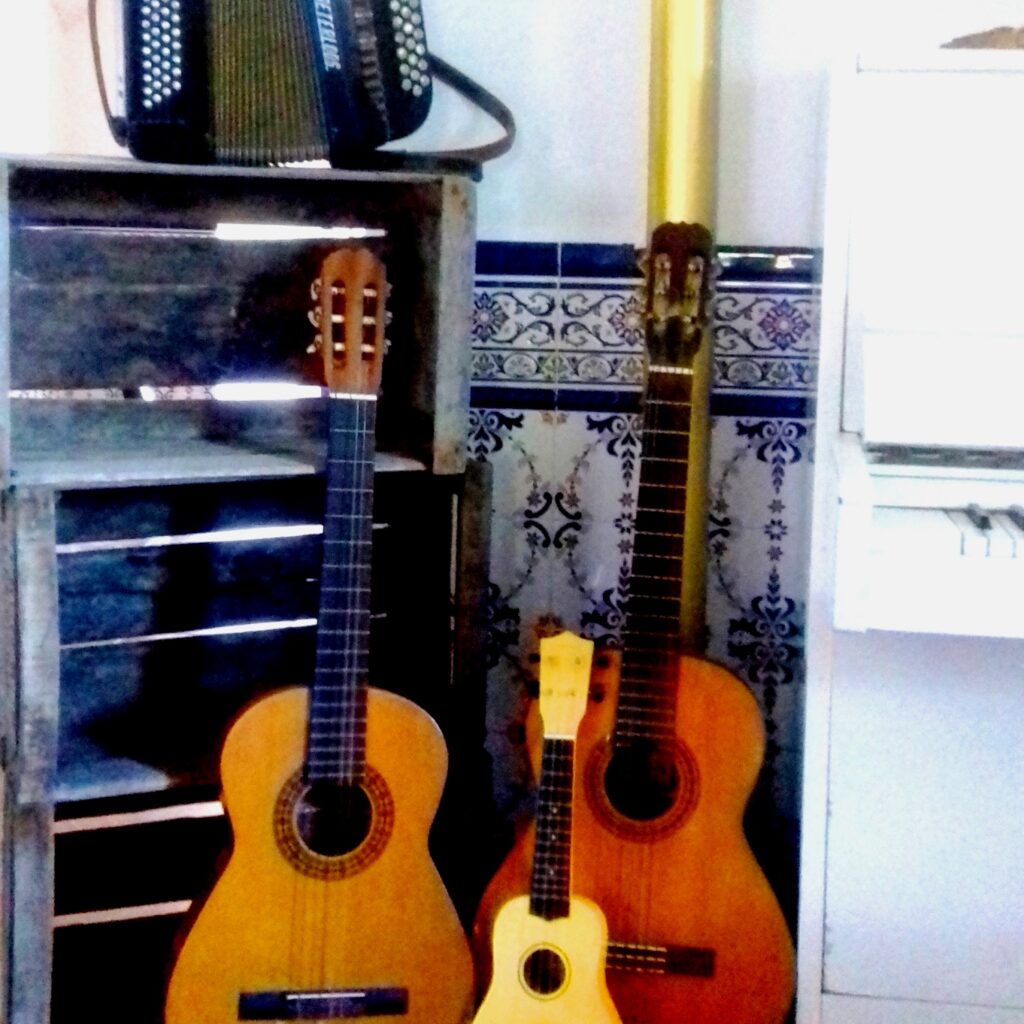 Zwei Gitarren und eine Ukulele lehnen an einem Regal aus Kisten, auf dem ein Akkordeon steht.