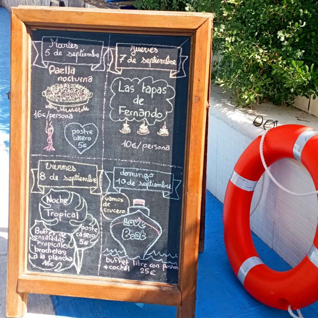 Grande ardoise de restaurant posée sur le sol d'une terrasse de piscine avec une bouée de sauvetage à droite. Sur l'ardoise est écrit à la craie en espagnol le programme de 4 soirées festives datées du 5 au 10 septembre