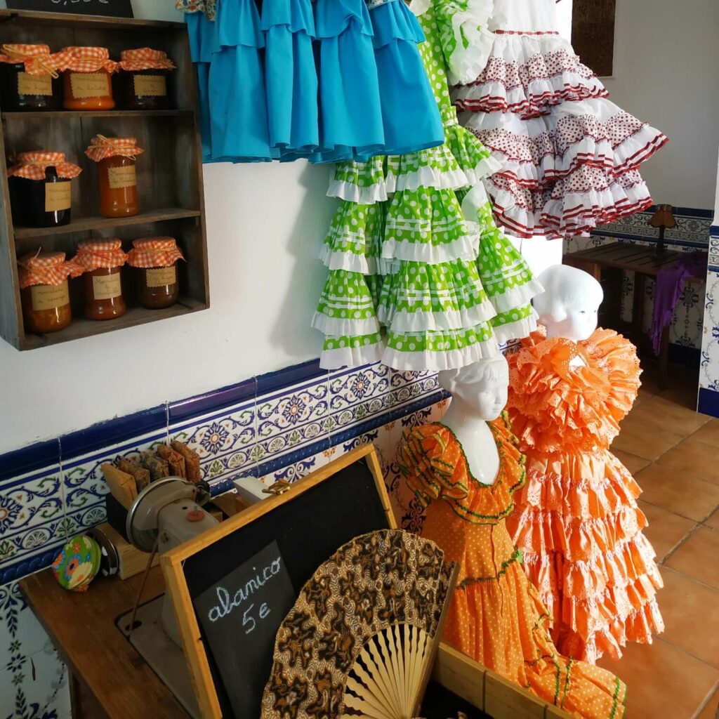 Interior of a shop with a small shelf of jam jars on the left, flamenco dresses for children on the right and a fan in the foreground.
